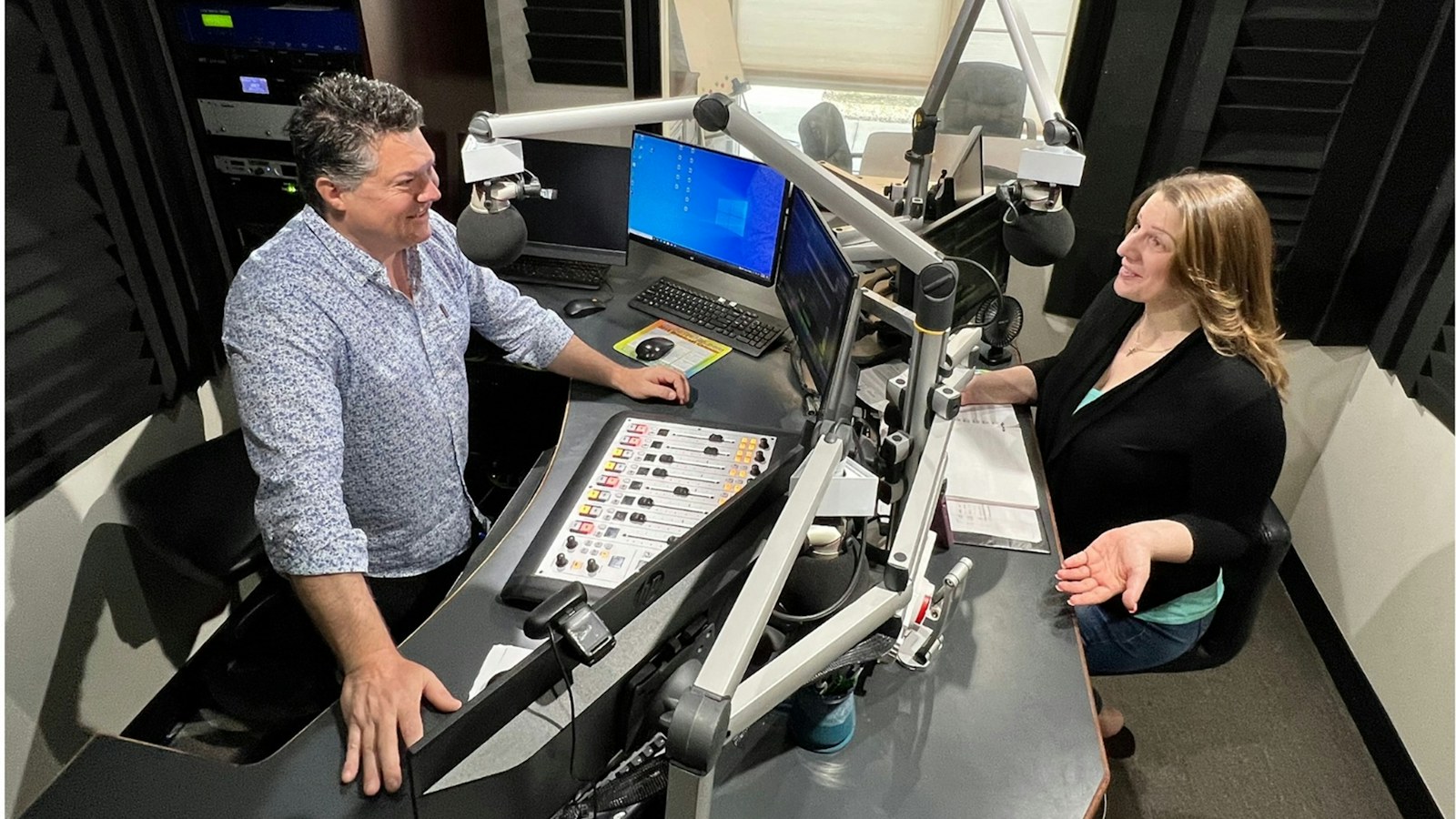 Two broadcasters talking across a radio studio console with the on-air board in front of them, the room where a radio format clock gets executed every hour
