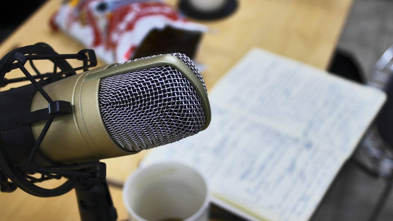 A gold studio microphone in a shock mount beside a handwritten page of show notes and a coffee cup, the desk where radio show ideas actually get worked out