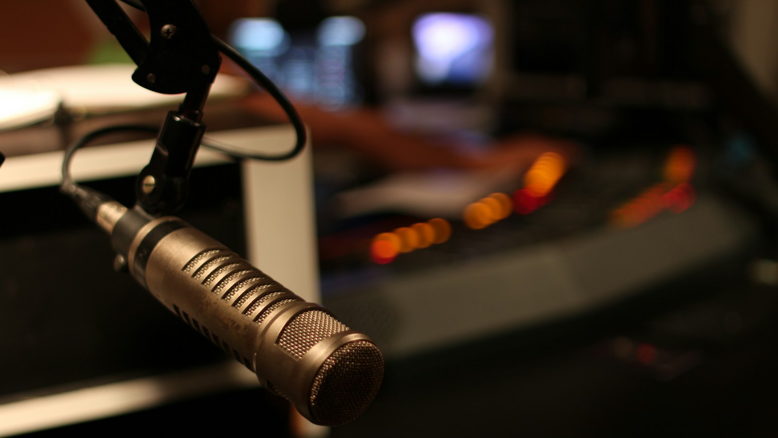 A broadcast microphone on a boom arm in a dim radio studio with a lit console and a stack of paper on the desk behind it, the place show prep actually gets used